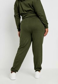 Set di abbigliamento da lounge a coste verde oliva, composto da una felpa con cappuccio e pantaloni jogger a gamba affusolata. Texture liscia, aderente, con polsini elasticizzati. Sneakers bianche.
