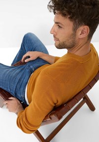 Senfgelber Pullover aus Strick mit gerippter Textur, Rundhalsausschnitt und langen Ärmeln, kombiniert mit blauen Jeans, sitzt auf einem Holzstuhl.