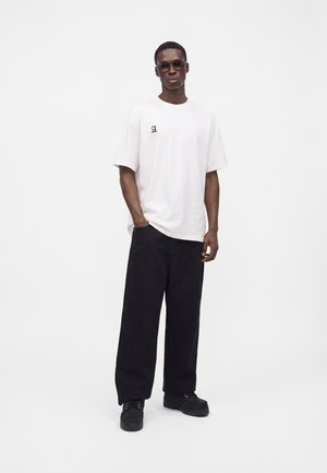 Muž so slnečnými okuliarmi, bielym oversized tričkom s malým čiernym písmenom, voľnými čiernymi nohavicami a čiernymi topánkami, stojaci na bielom pozadí.