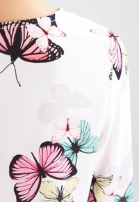 Tissu blanc avec un motif de papillons en rose, noir, turquoise et jaune. Dispose d'une texture douce, d'un léger tombé et d'un col rond.