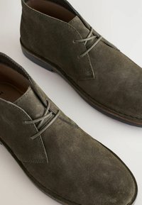 Botines de ante verde hasta el tobillo con una textura suave, que presentan una puntera redonda, costuras mínimas y detalles de cordones cruzados.