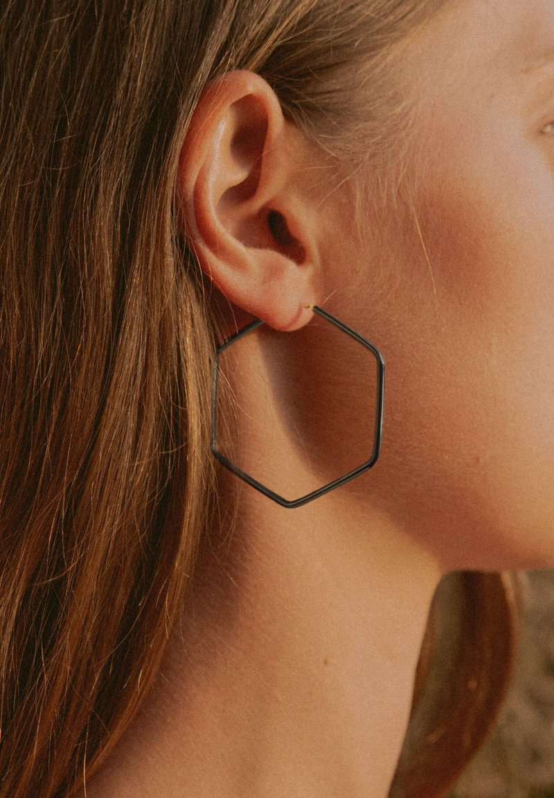 Schwarze Metall-Hexa-Ohrringe mit dünnem, minimalistischem Design. Zarte Glanzoberfläche, leicht und hängen an einem kleinen goldfarbenen Stecker.