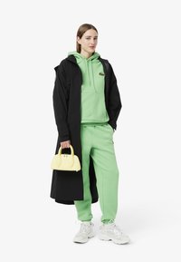 Felpa verde con cappuccio e pantaloni della tuta, cappotto lungo nero, sneakers bianche e una borsa gialla. Presenta un logo di coccodrillo sulla felpa.