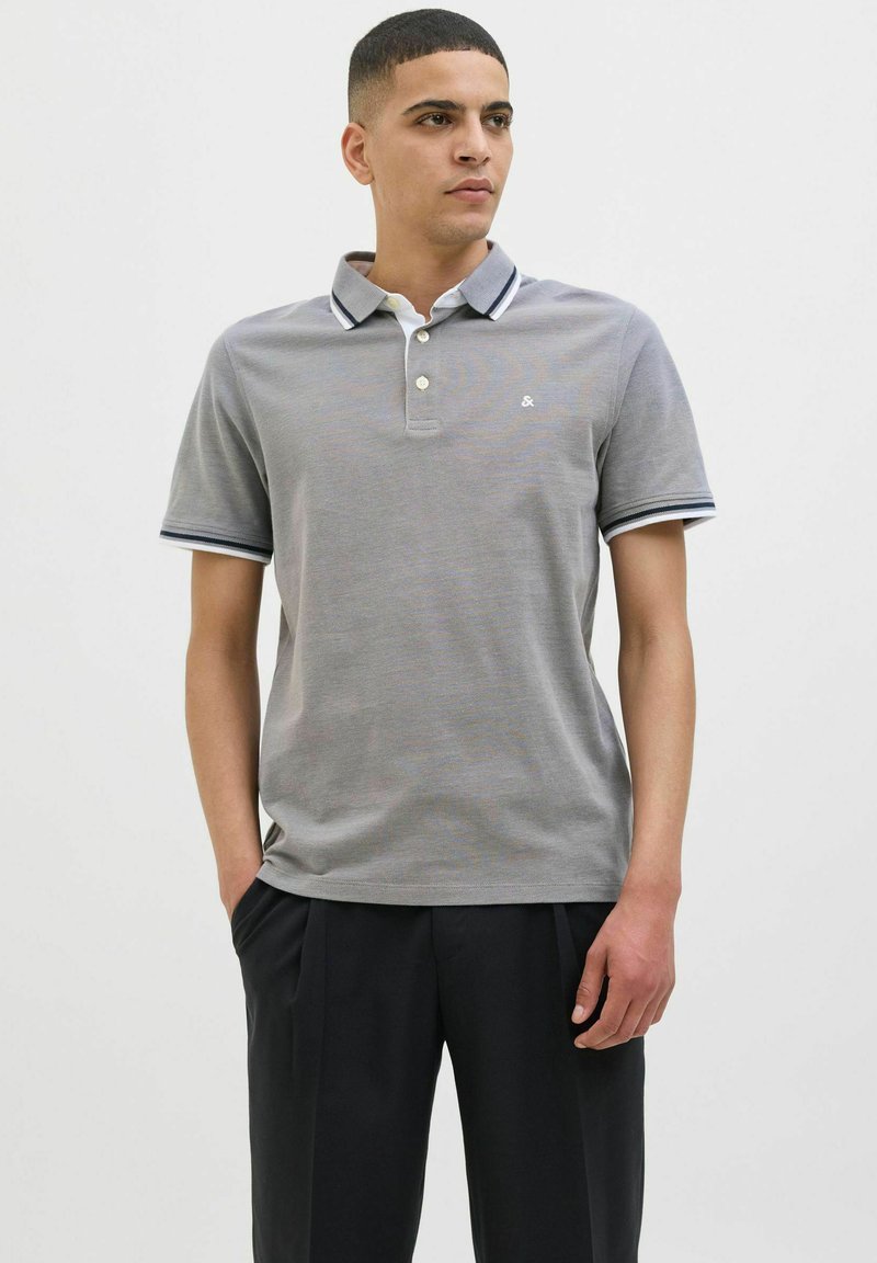 Grijze polo met een kraag, drie knopen en contrasterende donkerblauwe en witte strepen op de mouwen. Zachte textuur.