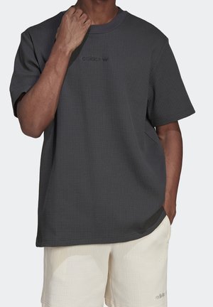 Man med grått strukturerad Adidas t-shirt och ljusbeiga strukturerade shorts, med ena handen som rör vid nacken och den andra handen i fickan.