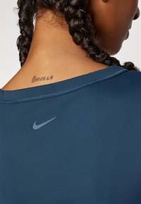 Top desportivo azul da Nike com decote redondo, mangas curtas e um logótipo discreto impresso no ombro esquerdo; apresenta uma tatuagem no pescoço.