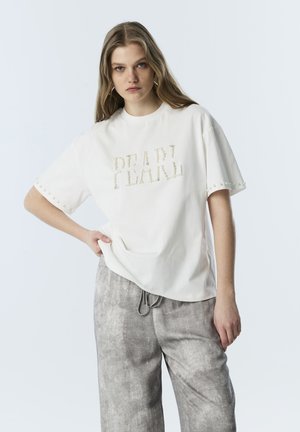 Joven mujer con cabello largo lleva una camiseta blanca de gran tamaño con letras de perlas y pantalones grises con cordón, de pie frente a un fondo liso.