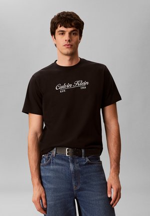 Młody mężczyzna ubrany w czarną koszulkę Calvin Klein z białym logo, niebieskie dżinsy i czarny pasek, stoi na jednolitym szarym tle.