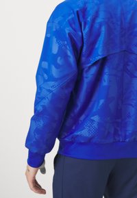 Chaqueta cortavientos azul con estampado, textura suave, cinturilla elástica y diseño de canesú trasero. Cuello alto y puños acanalados.