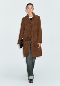 Bruine suede jas met een kraag, knopen aan de voorkant en zijzakken, gecombineerd met een donkere spijkerbroek en een zwarte clutch.