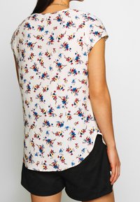 Blommig kortärmad blus i lätt tyg, med en krämfärgad botten och blå, röda och gula blommor, rundad nederkant och sidoklappar.