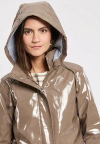 Giacca da pioggia lucida marrone chiaro con cappuccio, dotata di chiusura con zip, tasche frontali e un morbido rivestimento grigio.