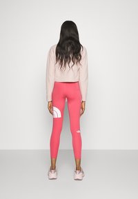 Tajta rosa leggings med en vit logotypmönster, matchade med en beige kortärmad sweatshirt. Modellen bär ljusrosa sneakers.