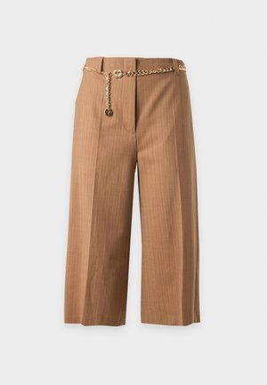 Femme portant un blazer beige à rayures fines avec un short large court assorti, un top court noir boutonné et des chaussures blanches à talons hauts.