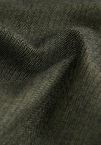 Tæt på mørkegrøn herringbone-mønstret stof med synlig tekstur og bløde folder, der skaber bløde skygger.