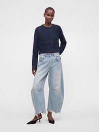 Personne portant un pull en maille duveteuse bleu marine foncé boutonné, un jean large déchiré bleu clair et des talons noirs pointus, debout devant un fond uni.