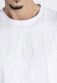Weißes Baumwoll-T-Shirt mit großen, halbtransparenten Buchstaben, die "KAVA" in einem Gittermuster bilden. Rundhalsausschnitt und kurze Ärmel.