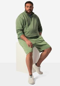 Grön tröja med kvartdragkedja, matchande gröna shorts och vita sneakers med svarta detaljer, sittande på en vit pelare.