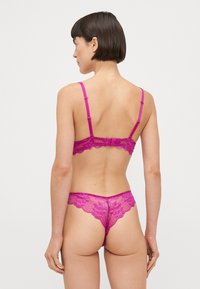 Ensemble en dentelle fuchsia comprenant un soutien-gorge et un string, avec des bords festonnés, des bretelles fines et réglables, et un design transparent avec un motif en grille sur le tissu.