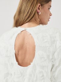 Vêtement blanc avec un détail texturé et des franges. Présente une grande découpe ovale à l'arrière et une fermeture à bouton au cou.