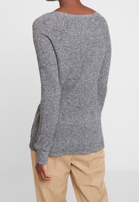 Grauer, gestrickter Pullover mit strukturierter Oberfläche, weitem Ausschnitt und langen Ärmeln, kombiniert mit beigen Hosen. Seitenschlitz für eine bessere Bewegungsfreiheit.