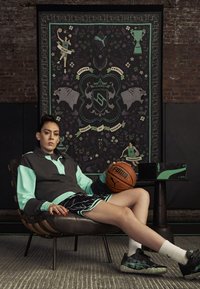 Atletska obleka s črno in metinjo barvno shemo; vključuje pulover, kratke hlače in pomlajen sneakers z vzorcem. Drži košarko ob vzorčenem ozadju.