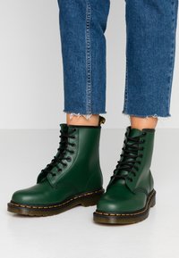 Green leather combat boots with a smooth finish, black laces, yellow stitching along the sole, and a reinforced toe.