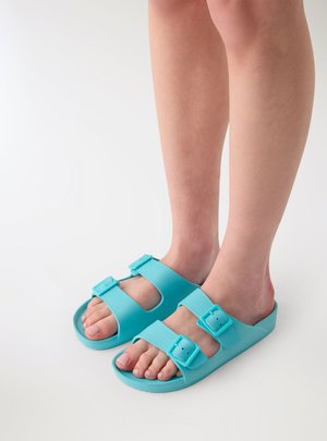Piedi che indossano sandali azzurri chiari con punta aperta e due cinturini regolabili con fibbia su sfondo bianco.