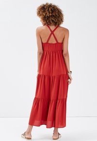 Robe maxi rouge rouille, avec bretelles croisées ajustables et volants à étages. Confectionnée dans un tissu léger à texture douce.