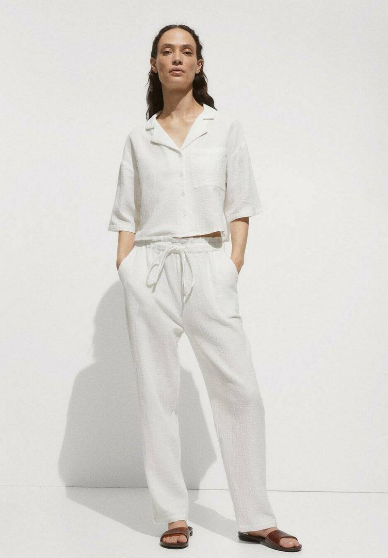 Witte, gemêleerde, shirt met korte mouwen en een zak, gecombineerd met witte, losvallende elastische broek. Bruine sandalen maken de outfit compleet.