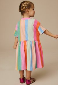 Multikleurige gestreepte jurk met korte mouwen, in roze, blauw, groen, oranje en geel. Lichte, geplooide rok. Roze sandalen.