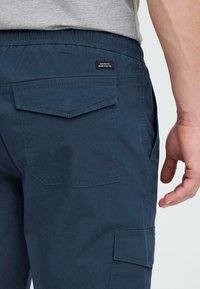 Donkerblauwe cargobroek met een elastische tailleband, zijzakken en twee achterzakken met een patch. De stof lijkt glad met minimale textuur.