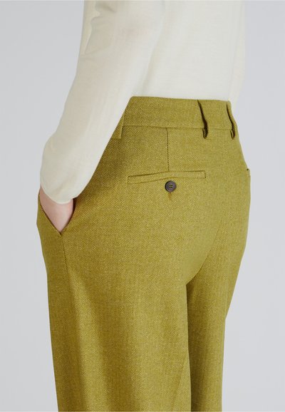 Persoon in lichtgele herringbone-patroon op maat gemaakte broek met handen in de zakken en een crèmekleurig langemouwen shirt.