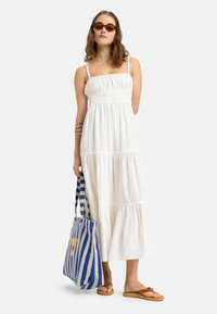 Witte maxi-jurk met een gerimpelde bodice, gelaagde rok en dunne schouderbandjes. Een blauwe en witte gestreepte tas met gele print en bruine sandalen.