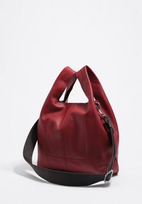 Raudona odinė rankinė su minkšta struktūra, dvigubomis rankenomis ir nuimamu tamsiu dirželiu. Žymosi užtrauktuko uždarymas ir subtilus siūlės detalizavimas.