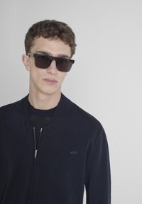Mornarska zip-up jakna s rebrastim ovratnikom, s blagim logotipom. Model nosi tamne sunčane naočale i crnu majicu s okruglim izrezom ispod.