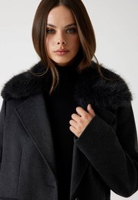 Palton din lână gri închis, cu guler din blană artificială neagră, rever larg și mâneci consistente. Paltonul are un design structurat, cu buzunare laterale.