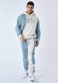 Sudadera color azul claro y crema con bloques de color, bolsillo delantero y texto bordado, combinada con joggers a juego que presentan un esquema y acentos de color similares.