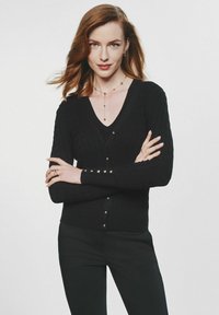 Czarny, prążkowany kardigan z dekoltem w serek i zapięciem na guziki. Ma długie rękawy z dodatkowymi guzikami. Nosi się go na czarną bluzkę.