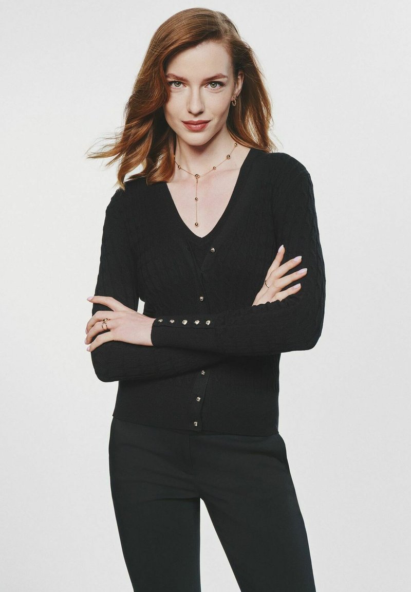 Czarny, prążkowany kardigan z dekoltem w serek i zapięciem na guziki. Ma długie rękawy z dodatkowymi guzikami. Nosi się go na czarną bluzkę.