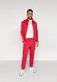 Κόκκινο αθλητικό σετ με λευκές ρίγες που περιλαμβάνει ζακέτα με φερμουάρ και παντελόνι που ταιριάζει. Περιλαμβάνει μπεζ μπλουζάκι και γκρι αθλητικά παπούτσια.