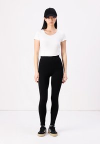 Zwarte high-waisted leggings gecombineerd met een witte shirt met korte mouwen en een zwarte pet, aangevuld met zwarte sneakers met een rubberen zool.