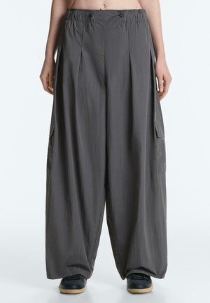 Személy laza, sötétszürke, bőszárú cargo nadrágot visel rugalmas derékrésszel és fekete cipőket gumitalppal.