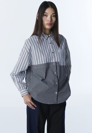 Junge Frau mit glatten dunklen Haaren, die ein lockeres, schwarz-weiß vertikal gestreiftes Hemd mit farbigen Knöpfen und dunkle Hosen trägt.