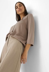 Donna appoggiata a una parete bianca, indossando pantaloni beige a gamba larga e un top taupe a maniche tre quarti, che guarda la fotocamera con sicurezza.