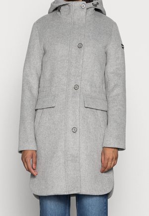 Persona que lleva un abrigo largo con capucha de color gris claro con botones y bolsillos con solapa, combinado con vaqueros azules.