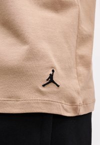 Μπεζ τελείωμα t-shirt με μαύρο κεντητό λογότυπο Jumpman πάνω από τη μαύρη μέση παντελονιού, δείχνοντας τη υφή του υφάσματος και τις λεπτομέρειες της ραφής.