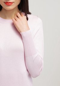Maglione rosa chiaro con scollo rotondo e polsi a coste, mostrato parzialmente sul corpo superiore di un modello, con texture a maglia morbida visibile.
