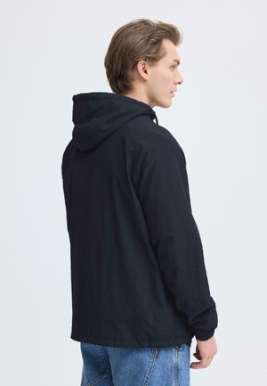 Man draagt een zwarte capuchonjas en blauwe spijkerbroek, kijkt naar rechts tegen een effen lichte achtergrond.