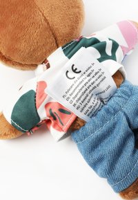 Plyšový medvěd hnědé barvy oblečený v barevné květinové košili a modrých džínových kalhotách, s etiketou péče na zádech. Celkově měkká textura.